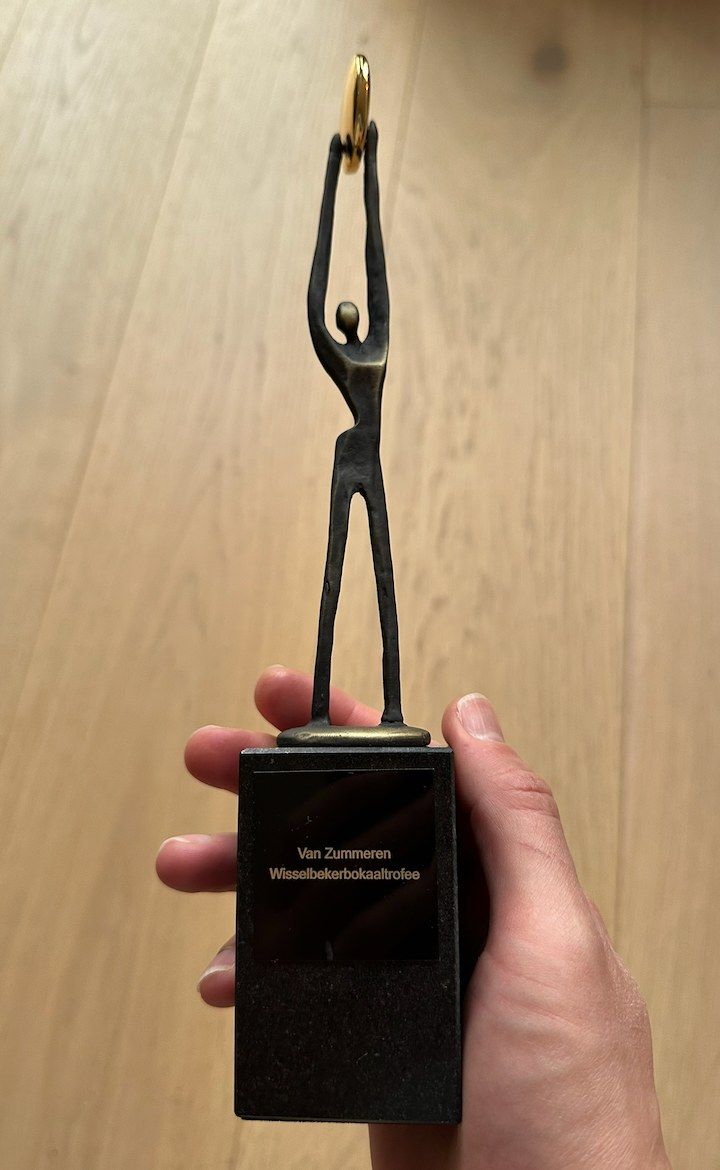 The real Van Zummeren trophy held in a hand: a thin bronze figure holding a golden ring above its head, on a black stone base with the name plate